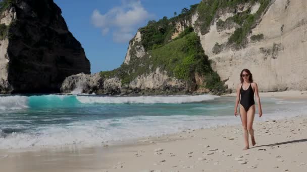 Brunett kvinna i svart baddräkt promenader längs havet stranden med stenar och turkos hav, blå himmel. Stranden Atuh, ön Nusa Penida, Bali, Indonesien. Tropiskt resekoncept. — Stockvideo