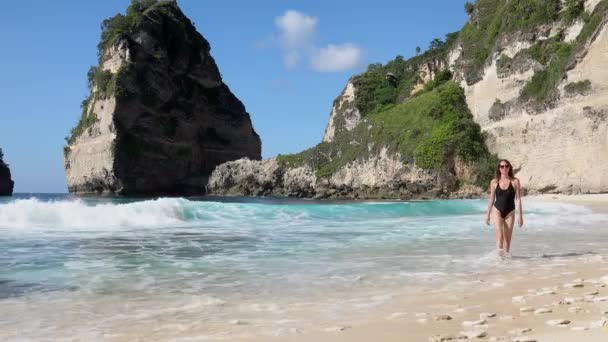 海の岩やターコイズブルーの海、青い空とビーチを歩く黒い水着のブルネットの女性。オーストラリアのビーチ、ヌサペニダ島、バリ、インドネシア。熱帯の背景と旅行の概念. — ストック動画
