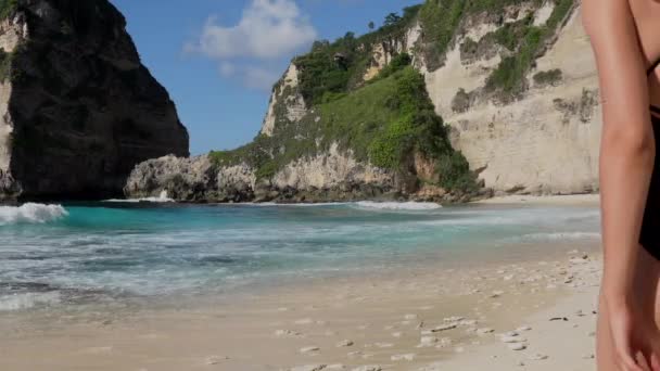 海の岩やターコイズブルーの海、青い空とビーチを楽しむ黒い水着のブルネットの女性。オーストラリアのビーチ、ヌサペニダ島、バリ、インドネシア。熱帯の背景と旅行の概念. — ストック動画