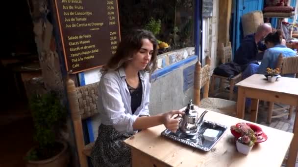 Marokko, Marrakesch - Oktober 2019: Eine europäische Touristin gießt und trinkt nach marokkanischer Tradition Minztee aus einer silbernen Teekanne ins Glas an einem kleinen Tisch. Rituelle Zubereitung von Tee — Stockvideo
