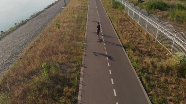 Pandangan udara manusia naik longboard di jalan lurus kosong di sisi sungai. Liburan musim panas naik skateboard di luar kota pada waktu matahari terbenam 4k — Stok Video
