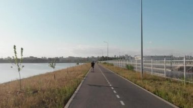 Nehir kenarındaki boş düz yolda uzun tahta süren bir adamın hava manzarası. Yaz tatili hippileri gün batımında şehir dışında kaykay sürerler.