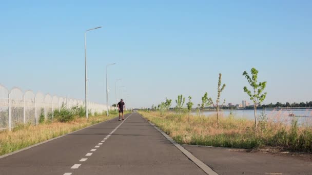 Vista frontal del hombre montando longboard en el camino recto vacío en el lado del río. Vacaciones de verano hipster en paseos sombrero monopatín fuera de la ciudad al atardecer 4k — Vídeos de Stock