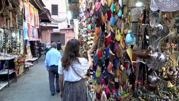 Marokko, Marrakech - oktober 2019: Traditionele Marokkaanse Marrakech bazaar. Europese vrouw kiezen Arabische handgemaakte leren schoenen in marktkraam met souvenirs en zilveren lantaarn decor. — Stockvideo