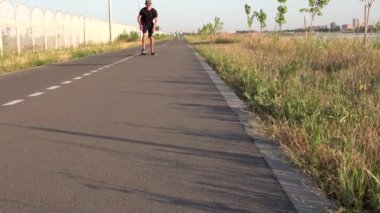 Boş düz yolda oturan bir adamın uzun tahta sürüşünü yakından görmek. Yaz tatili hipster 'ı gün batımında şehir dışında kaykaya biner. Yüksek kalite 4k görüntü