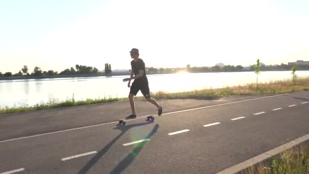 Hipster man rider longboard njuter cruising på väg nära floden stranden i solnedgången ljus, bär hatt, solglasögon. Sommar fritid aktivitet, träna utanför — Stockvideo