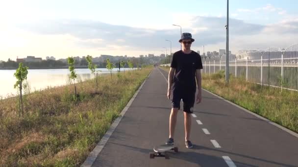 Hipster homem fica com longboard e começa a se mover, usando óculos de sol e chapéu ao pôr do sol tempo com horizonte da cidade. Conceito de lazer de verão. Desporto ao ar livre ativo. — Vídeo de Stock