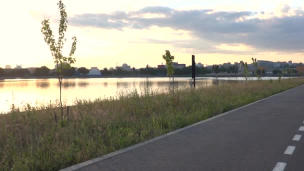Ensam longboard på asfalterad väg väg på flodstrand i solnedgången i park utanför staden med skyline — Stockvideo