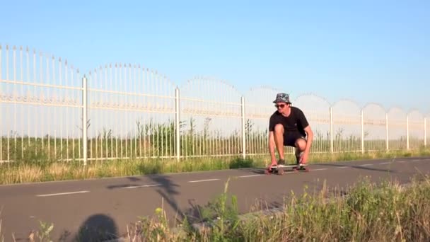 Homem monta longboard sentado na estrada reta vazia e tocar seus óculos de sol. Verão férias hipster passeios de skate fora da cidade ao pôr do sol. — Vídeo de Stock