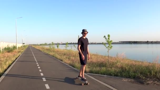 Fiatal hipszter férfi lovaglás longboard élvezi cirkáló gyönyörű vidéki úton, közel a folyóparton. A kamera közelről követi a fickót és elkezd gördeszkázni naplementekor.. — Stock videók