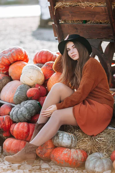 Portret van een vrolijke vrouw in de buurt van een wagen met sinaasappelpompoen op de boerenmarkt in bruine trui, jurk en hoed. Gezellige herfst vibes Halloween, Thanksgiving dag — Stockfoto