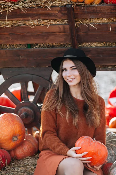 Портрет счастливой улыбающейся женщины, сидящей возле деревянного фургона с оранжевой тыквой на фермерском рынке в коричневом свитере, платье и шляпе. Уютные осенние вибрации Хэллоуин, День Благодарения — стоковое фото