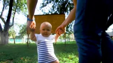 Küçük bebek yürümeyi öğreniyor. İlk Basamaklar. Ebeveynler çocuklarına yaz aylarında yeşil çimlerde ilk adımları atmayı öğretiyor. Mutlu çocukluk ve ebeveynlik kavramları. Aile sevgisi