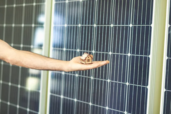 man holds on his hand a model of a wooden house on a background of solar panels
