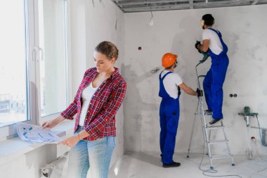 Bir bayan müşteri, arka plandaki tamircilerin bir matkapla merdiven üzerinde durarak bir daire planlamasını inceler.