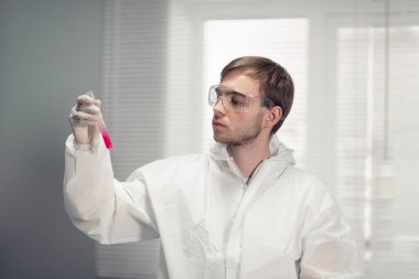 Koruyucu gözlüklü bir erkek doktor elinde beyaz bir şişe tutuyor.