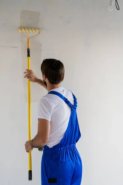 Homem de macacão azul pintando a parede com um rolo, vista da parte de trás — Fotografia de Stock