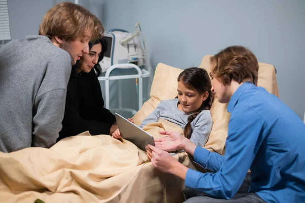 A sick kid in a hospital bed having visitors, the girl is listening to her brother attentively while he is teaching her to use a new app