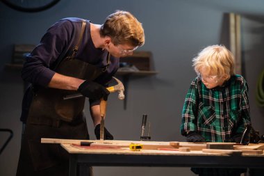 Genç bir erkek marangoz atölyesinde genç bir çocuğa marangozluk öğretiyor..