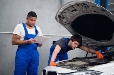 Bir adam bozuk bir arabayı inceler. Ortağı not alıyor.