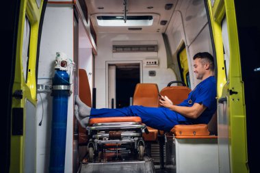Mavi üniformalı genç bir stajyer ambulans arabasında dinleniyor ve telefonunu karıştırıyor.