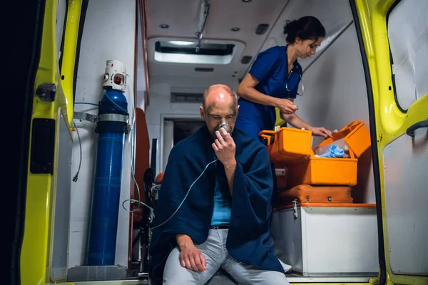 L'homme respire à travers le masque à oxygène, jeune infirmière emballe l'équipement dans un sac médical dans la voiture d'ambulance — Photo