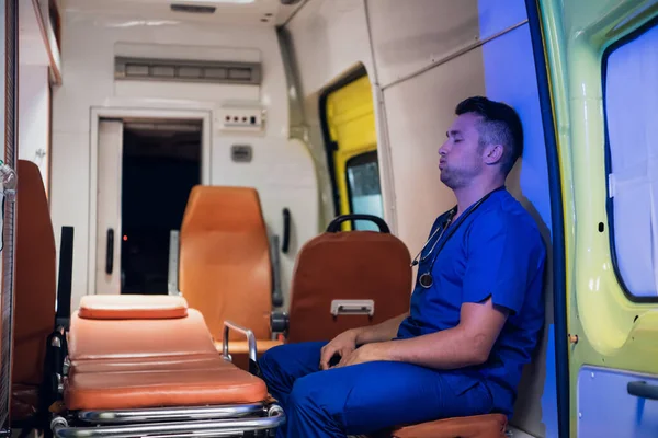 Парамедик в синей форме сидит в машине скорой помощи, делает глубокий вдох, отдыхает. — стоковое фото