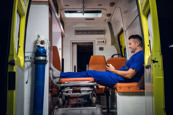 Junger Praktikant in blauer Uniform ruht sich aus und surft im Krankenwagen mit seinem Handy — Stockfoto