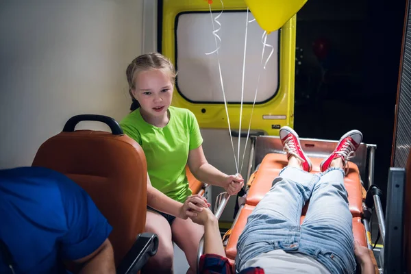 Грустная девушка смотрит на женщину, лежащую на носилках в машине скорой помощи, девушка держит ее сестер за руку — стоковое фото