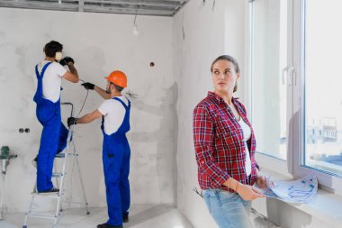 Müşteri pencerede durur ve dairenin düzenini inceler. Tamirciler arka planda çalışır.