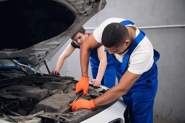 Two male mechanics in uniform inspect a car engine
