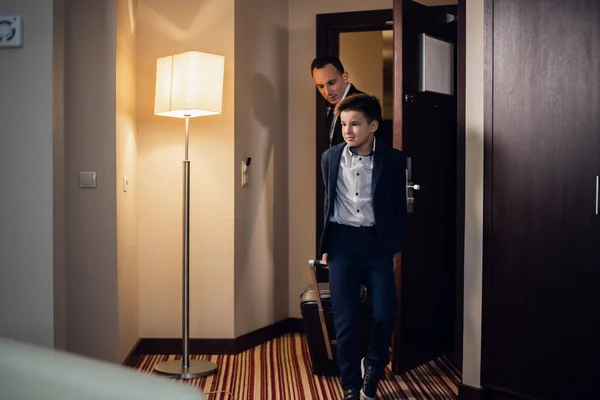 Father and son in formal wear entering a hotel room with suitcas