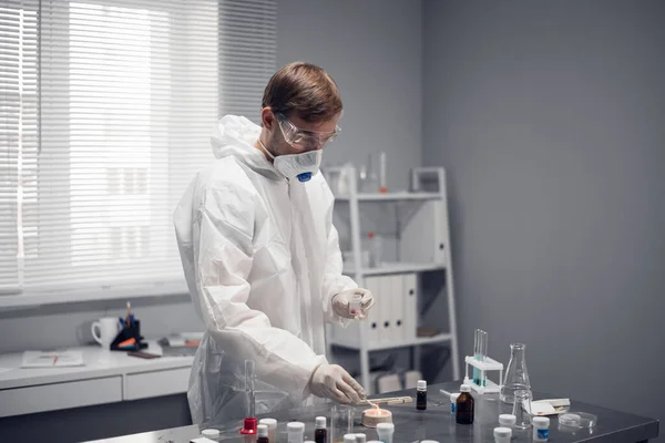 Um cientista em óculos de proteção, respirador e macacão branco em pé à mesa no laboratório de ciências, trabalhando em algo — Fotografia de Stock