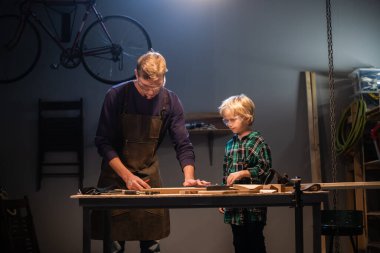 Genç bir erkek marangoz atölyesinde genç bir çocuğa marangozluk öğretiyor..