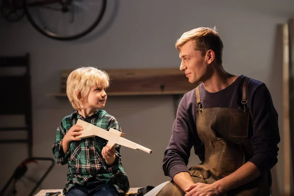 a young carpenter dad and his cute blond son made a gun out of wood and show it, they are happy, sitting in the carpenters workshop on the table.