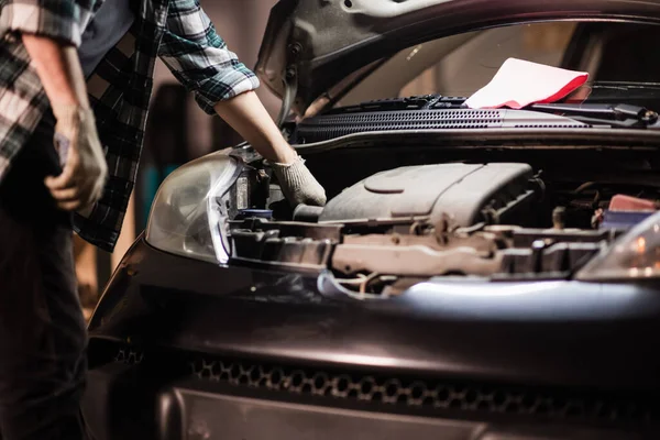 Młody mechanik samochodowy w koszuli i rękawiczkach sprawdza poziom oleju silnikowego w czarnym samochodzie. — Zdjęcie stockowe