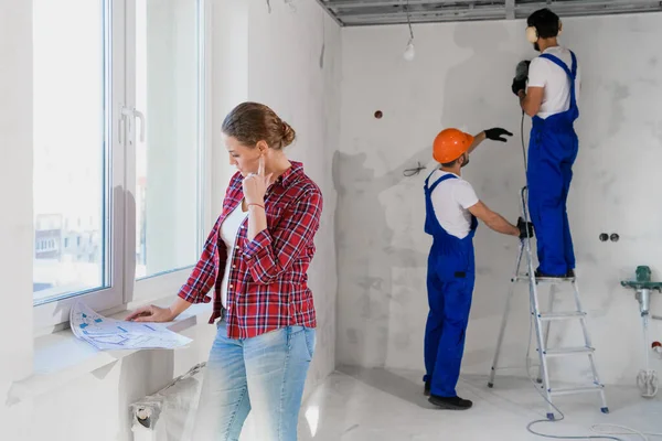 A nő megvizsgálja a lakás elrendezését, az építők egy létrán fúrják a falat. — Stock Fotó