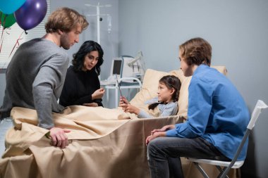 Hastane yatağında hasta bir çocuğun ziyaretçisi var. Kız elinde bir tablet tutarken annesini dikkatle dinliyor.