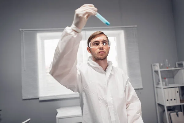 Retrato de close-up do cientista segurando um tubo com algum líquido azul contra a luz e olhando atentamente para ele — Fotografia de Stock