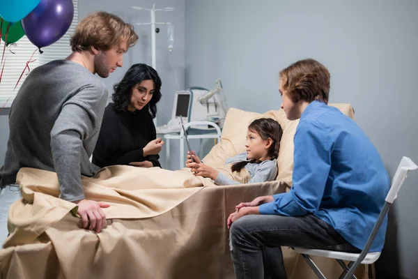 Hastane yatağında hasta bir çocuğun ziyaretçisi var. Kız elinde bir tablet tutarken annesini dikkatle dinliyor.