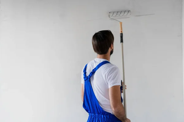파란 작업복을 입은 건축가가 로울러 로 벽을 칠하고, 뒤에서 바라본다 — 스톡 사진