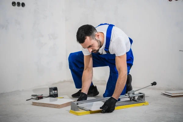 Ein Arbeiter in einem blauen Overall schneidet mit einem Cutter eine Fliese — Stockfoto