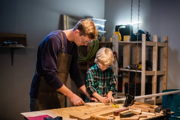 a young father and his little son make a toy out of wood in a carpenters workshop.
