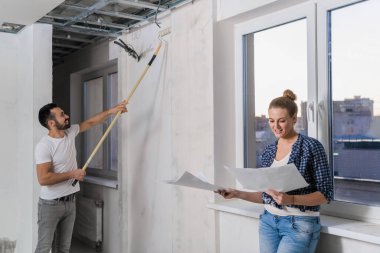 Bir adam silindirle duvarı boyuyor, bir kadın da dairenin düzenini inceliyor.