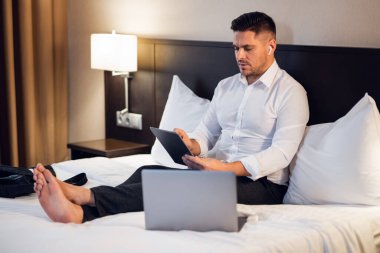 Tableti ve laptopuyla yatakta çekici bir iş adamı.