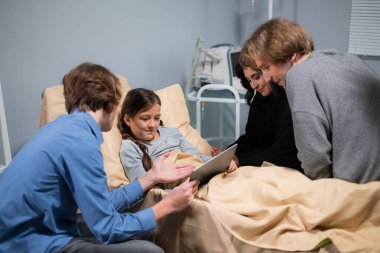 Küçük mutlu bir kız hastanede yatarken tablet kullanmayı öğreniyor. Ağabeyi de ona yardım ediyor.