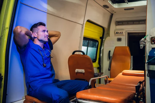 Санитар в синей форме сидит в машине скорой помощи с руками на затылке. — стоковое фото
