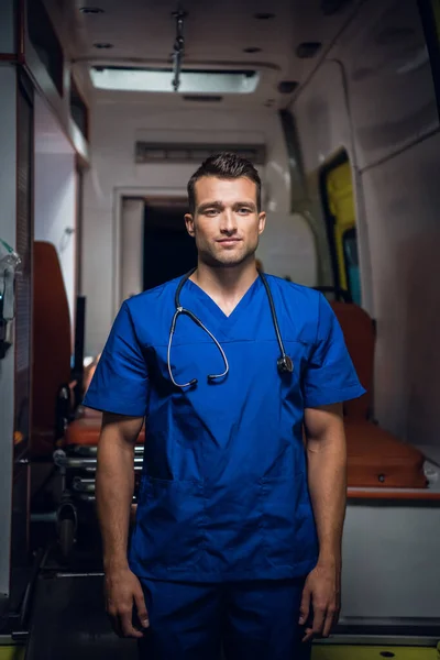 Un joven médico con uniforme azul se para y mira la cámara delante de un coche de ambulancia —  Fotos de Stock