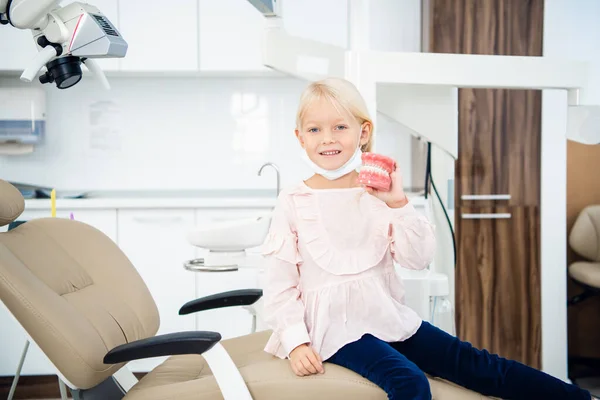 Una niña pequeña sostiene dentaduras artificiales en su mano en el consultorio dental — Foto de Stock