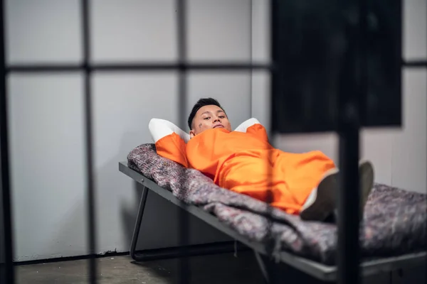 Een gevangene in een Aziatische gevangenis ligt op een bed in een rood gevangenenuniform in een eenzame cel.. — Stockfoto